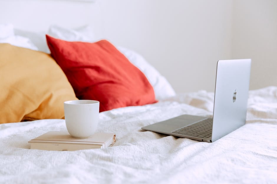 Laptop and cup on a bed with colorful pillows, evoking a modern cozy workspace.