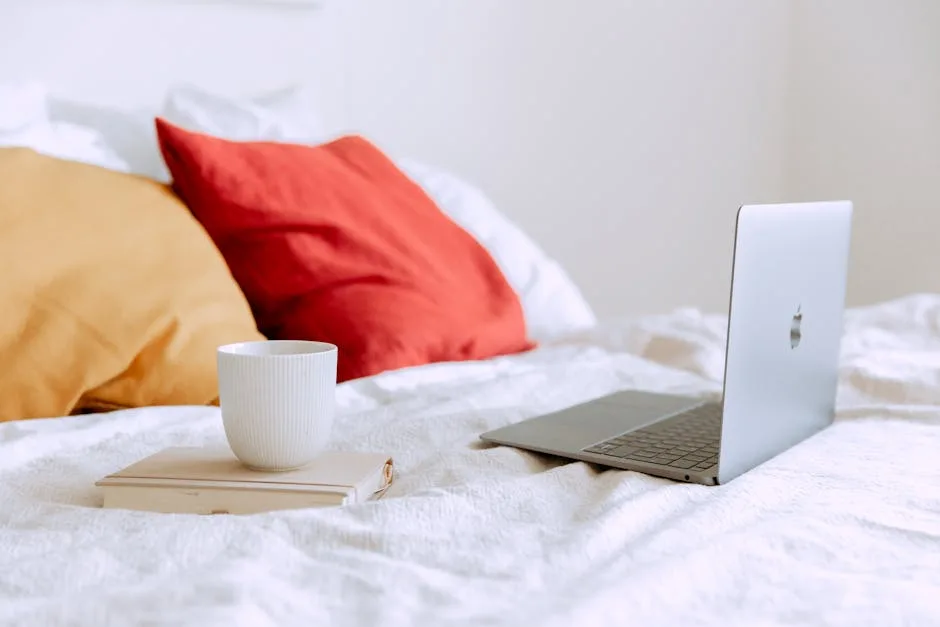 Laptop and cup on a bed with colorful pillows, evoking a modern cozy workspace.